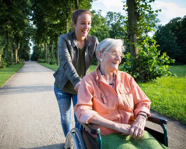 Eine jüngere Frau schiebt eine ältere Dame im Rollstuhl | © fotografixx - Getty Images/iStockphoto